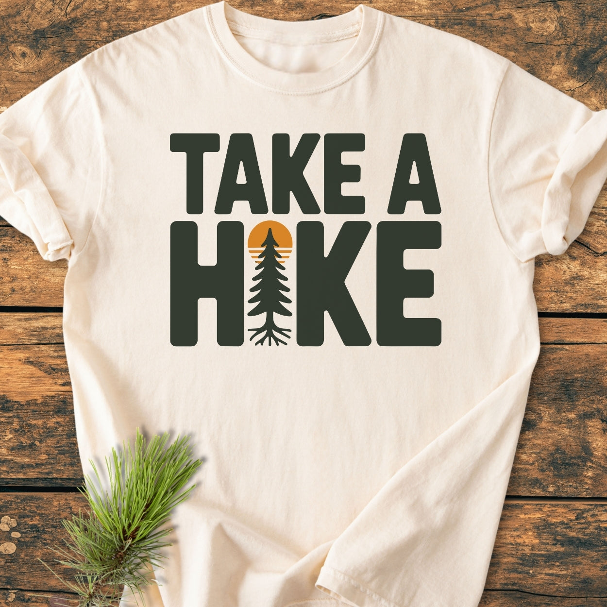 Take A Hike - Vintage Wash Tee on wooden surface, features bold green text with a pine tree in place of the I, sun detail. Pine sprig nearby enhances its adventurous theme.