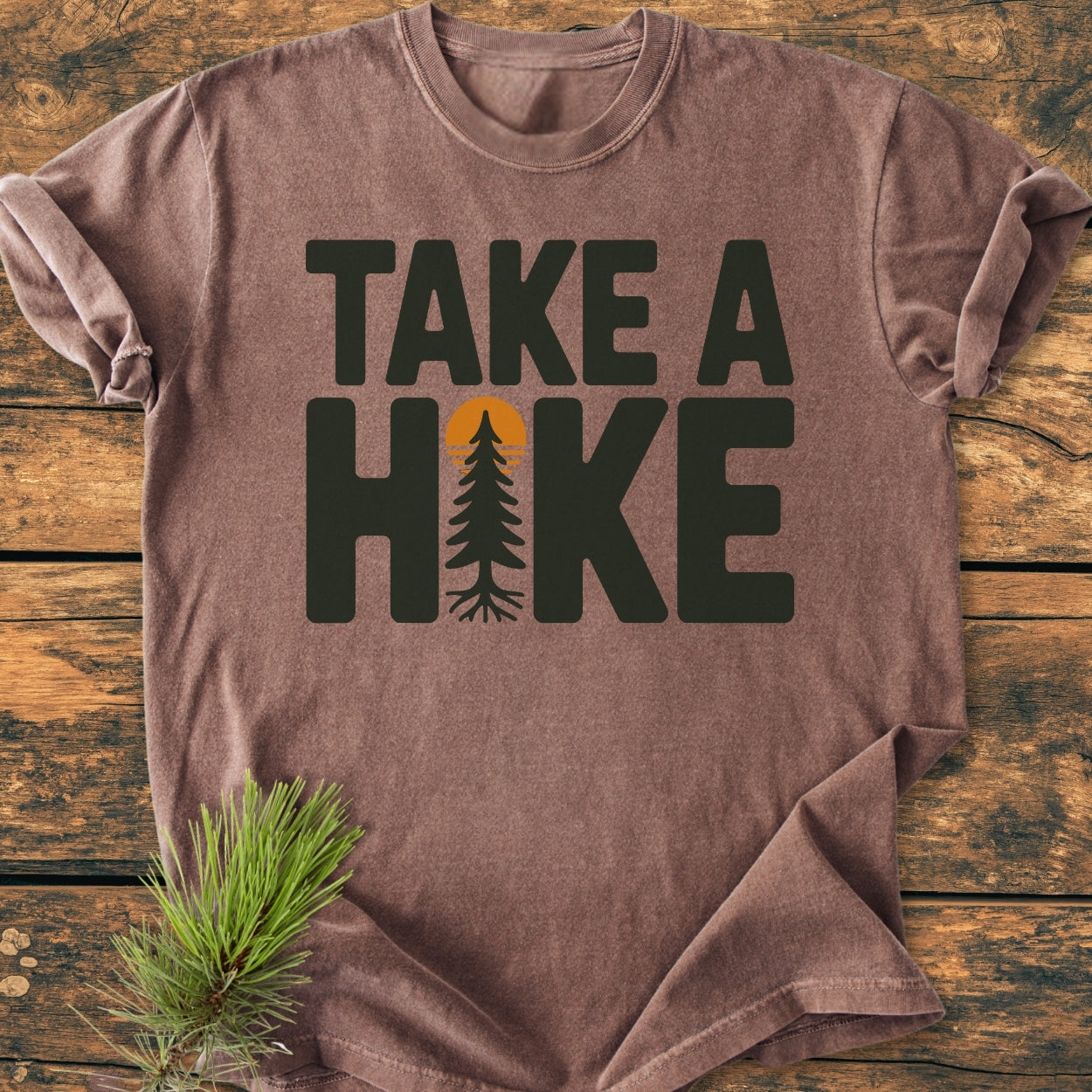 Take A Hike - Vintage Wash Tee displayed on wood, featuring a stylized pine tree as the letter I in HIKE with an orange sun, complemented by a pine needle sprig.
