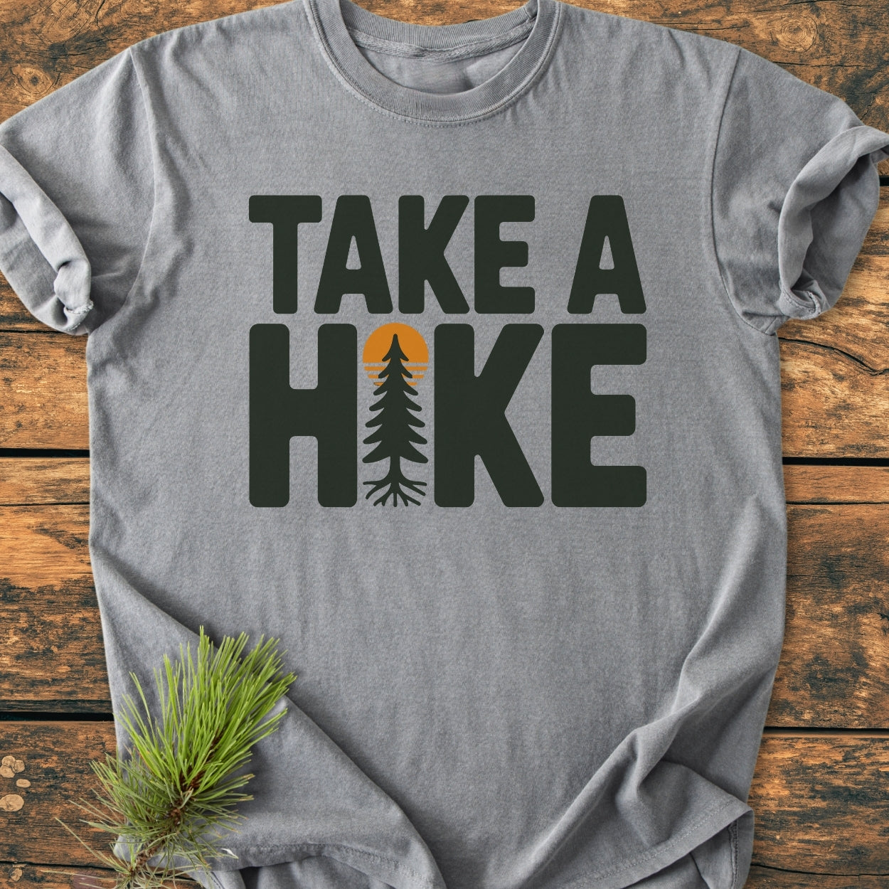 Take A Hike - Vintage Wash Tee: Heather-gray T-shirt with TAKE A HIKE in dark green and a pine tree design, laid flat with pine needles accent, emphasizing cozy, durable, relaxed fit.