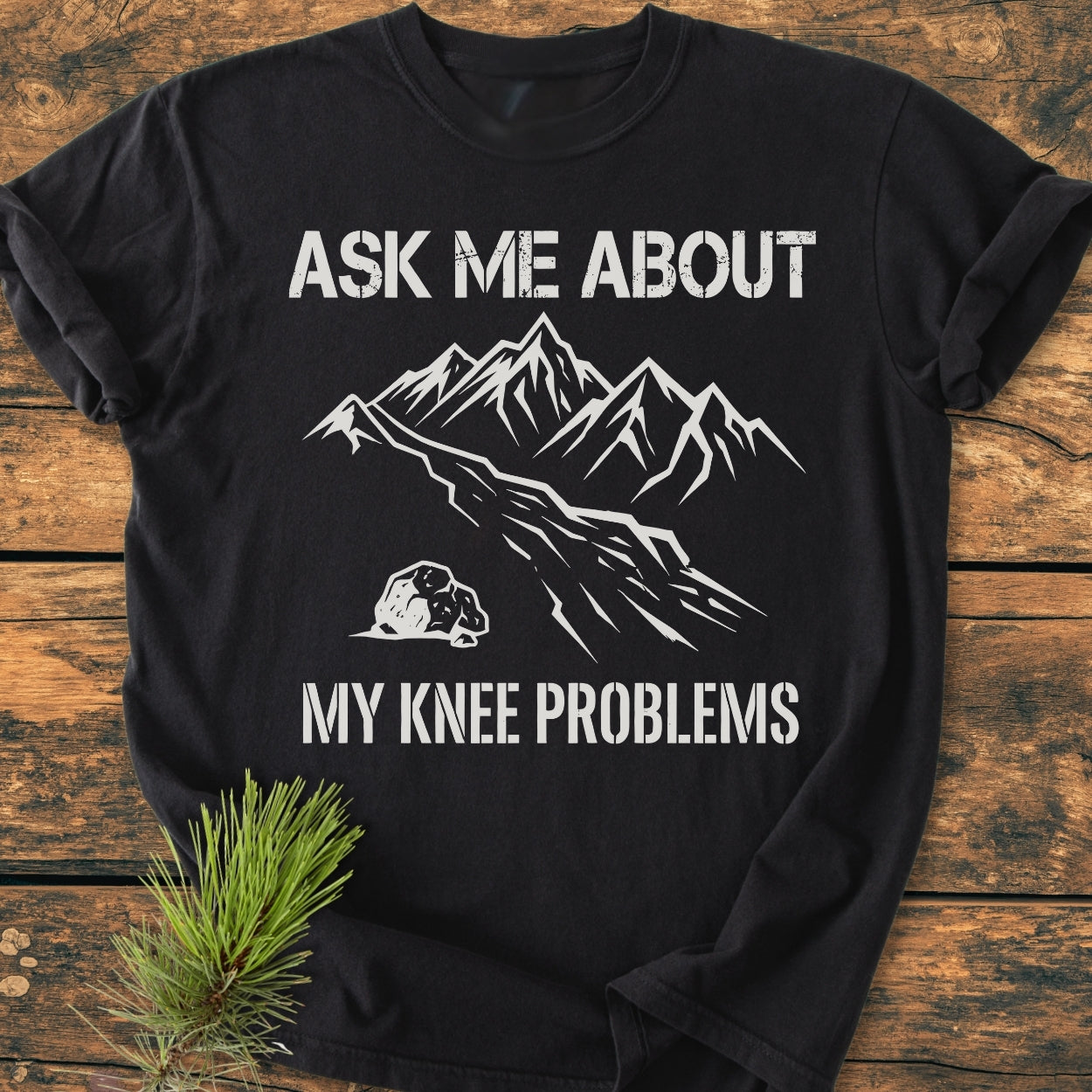 Ask Me About My Knee Problems - Vintage Wash Tee featuring a white mountain range design printed on a black T-shirt, laid on wood with a pine sprig beside it.