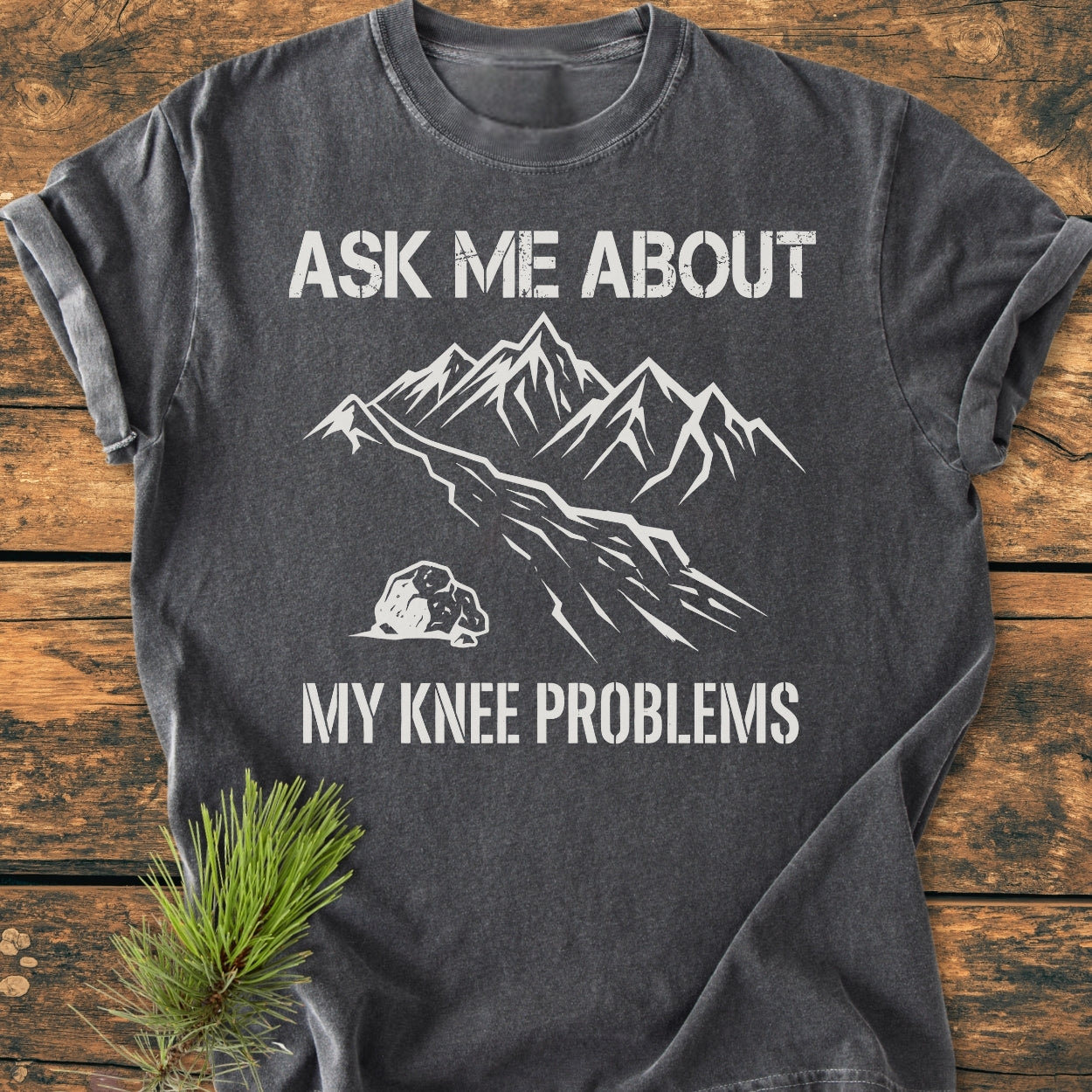 Ask Me About My Knee Problems - Vintage Wash Tee: Charcoal-gray shirt with bold white slogan and mountain graphic, laid on wood. Features soft, garment-dyed cotton for comfort and relaxed fit.
