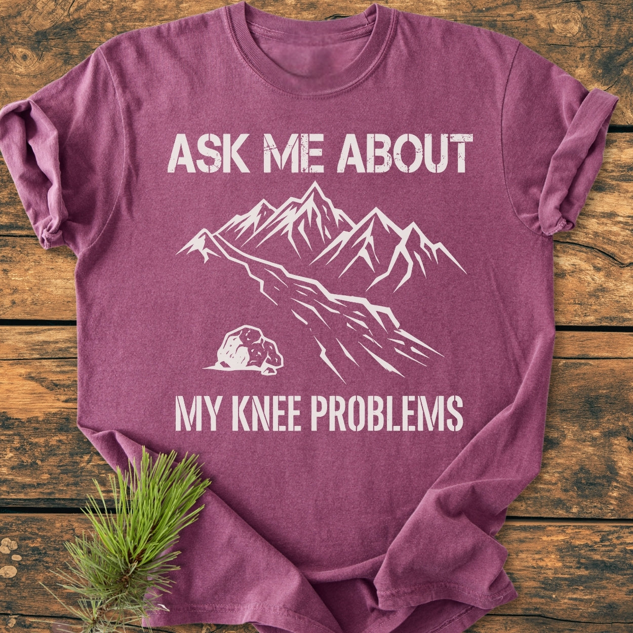 Ask Me About My Knee Problems - Vintage Wash Tee featuring a stylized mountain design on a mauve T-shirt, displayed on wood with pine needles, highlighting its relaxed fit and durable stitching.