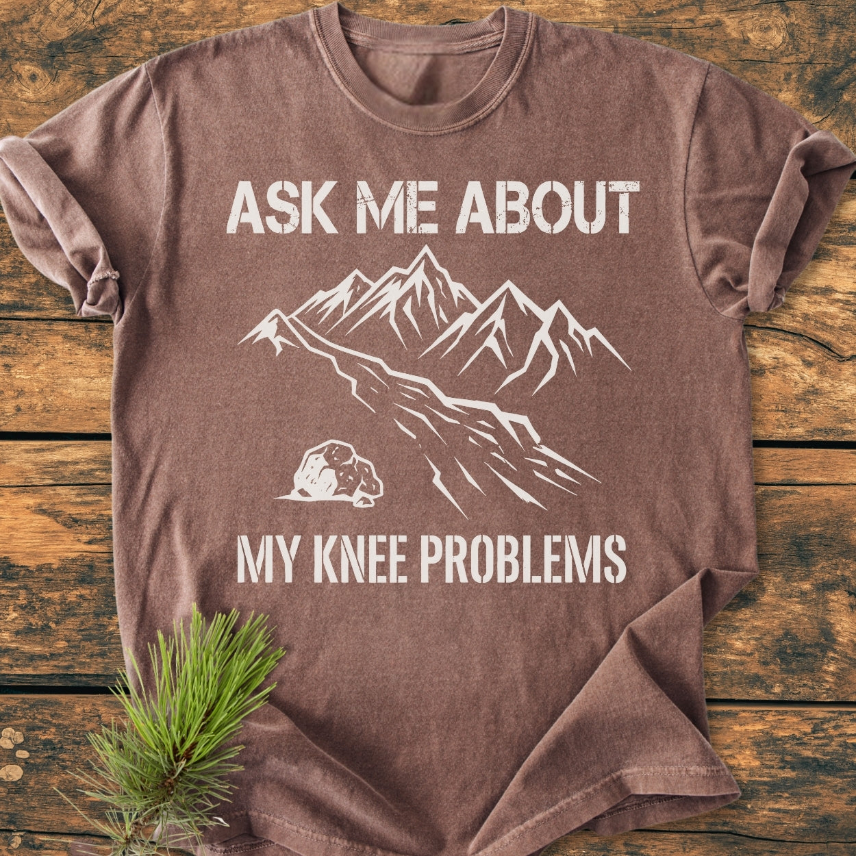 Ask Me About My Knee Problems - Vintage Wash Tee, shown on a wooden surface, features block letters and a mountain graphic. Made from soft, durable ring-spun cotton, offering a relaxed fit.
