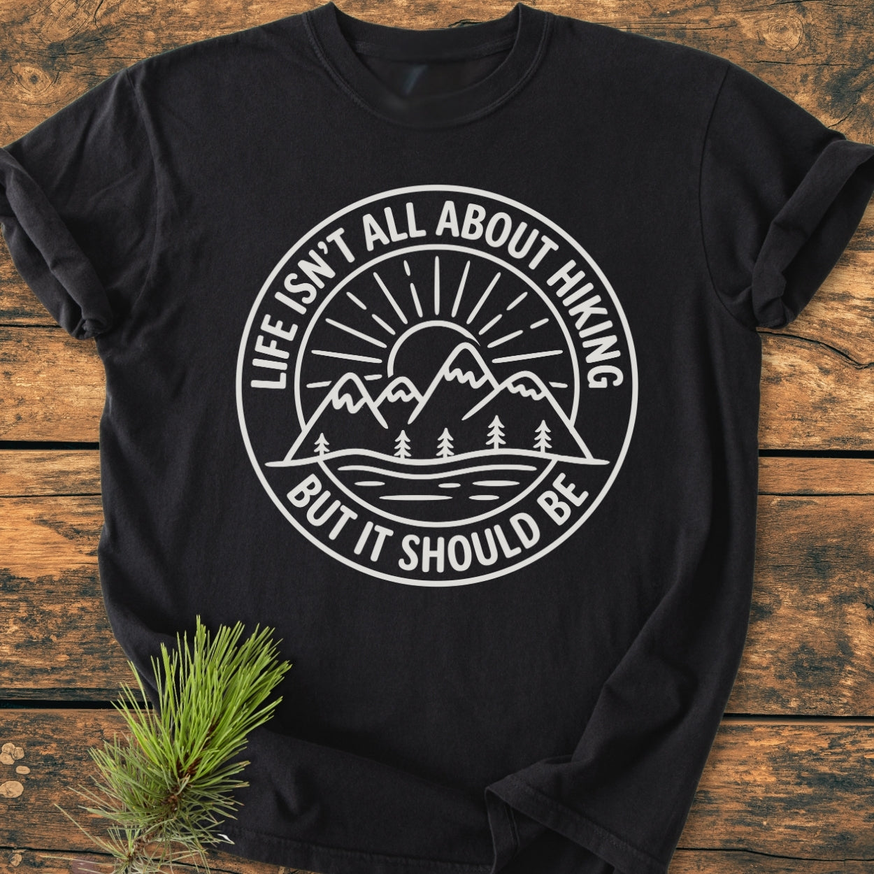 Life Isn't All About Hiking - Vintage Wash Tee on rustic wood, featuring a white emblem of mountains, sunburst, trees, and water, with bold slogan emphasizing hiking passion. Pine sprig accent nearby.