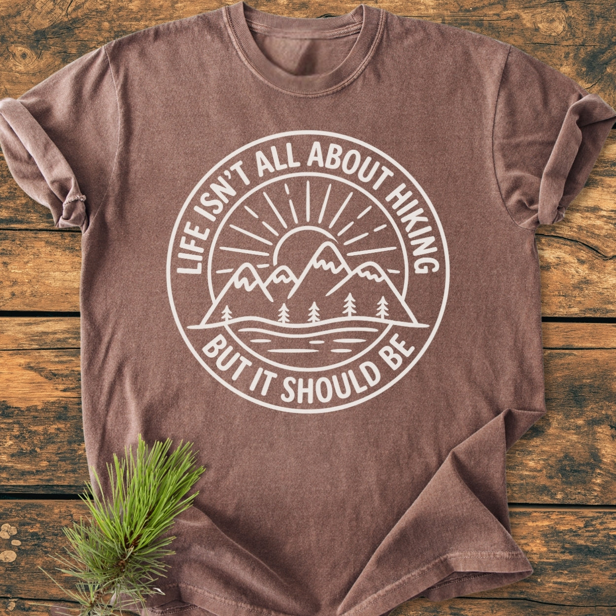 Life Isn't All About Hiking - Vintage Wash Tee features a white sunrise graphic over mountains. Laid flat on wood, it embodies comfort with garment-dyed, 100% ring-spun cotton for a relaxed fit.