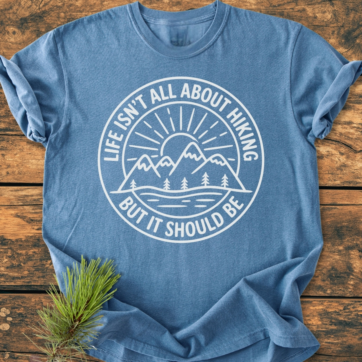 Life Isn't All About Hiking - Vintage Wash Tee features a white graphic of mountains, trees, sun, and river on a blue heather-style shirt, emphasizing outdoor adventure, with a spruce branch accent.