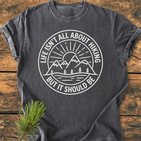 Life Isn't All About Hiking - Vintage Wash Tee features a white graphic of mountains and a sun on dark-gray fabric, laid flat on wood, with a pine bough accent.
