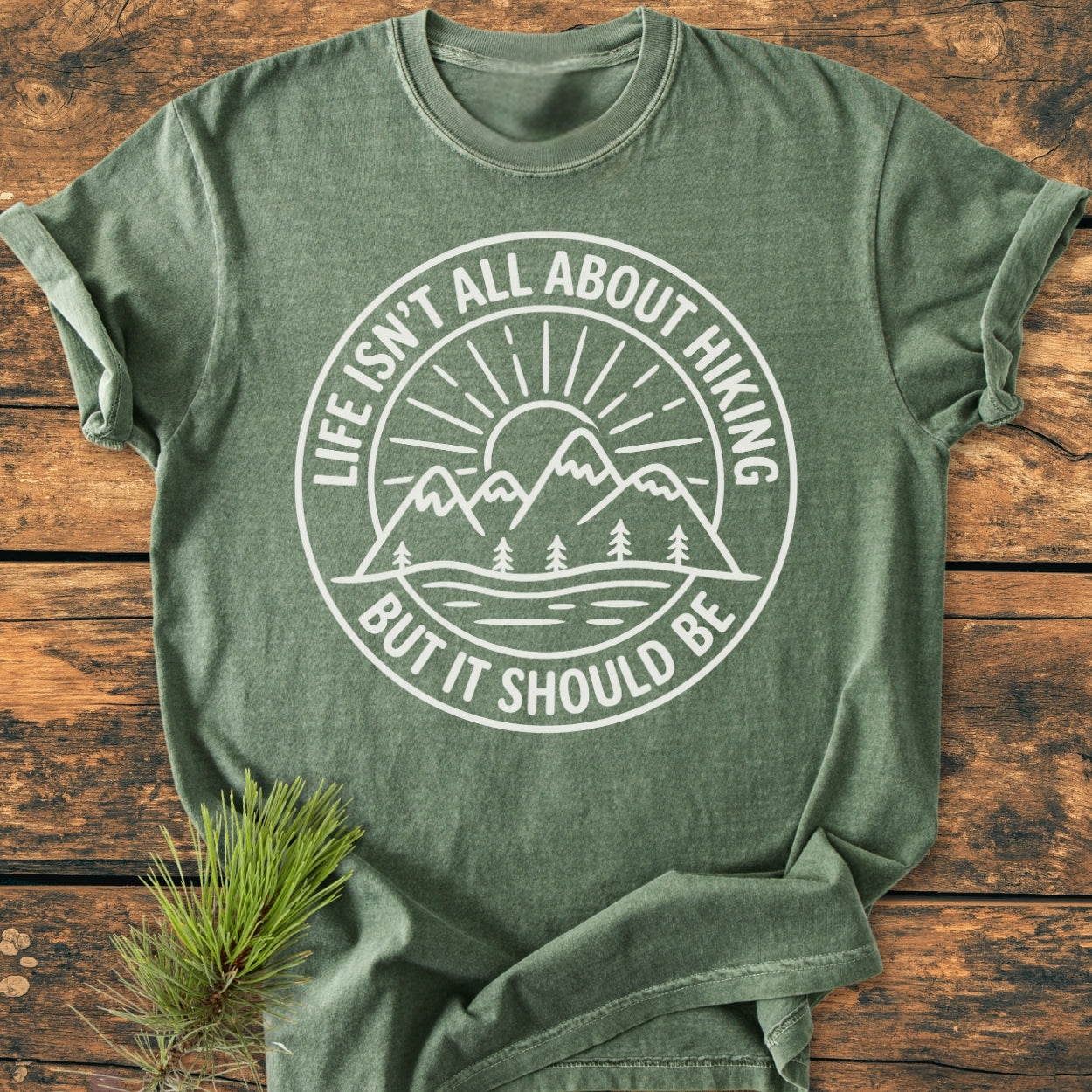 Life Isn't All About Hiking - Vintage Wash Tee features a circular emblem with mountains, trees, sun, and water, screen-printed in white on a heather-green T-shirt, laid on rustic wood.