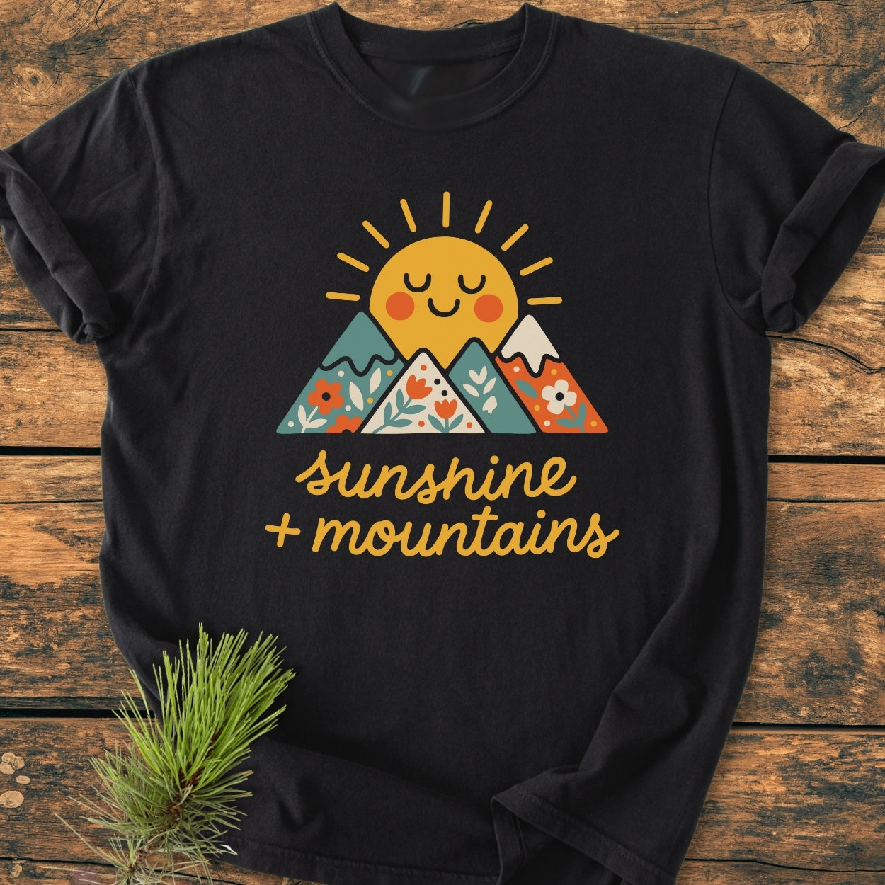Sunshine And Mountains - Vintage Wash Tee features a smiling sun and flower-patterned mountains graphic. Laid on wood with a pine sprig, showcasing its relaxed fit and soft, garment-dyed cotton fabric.