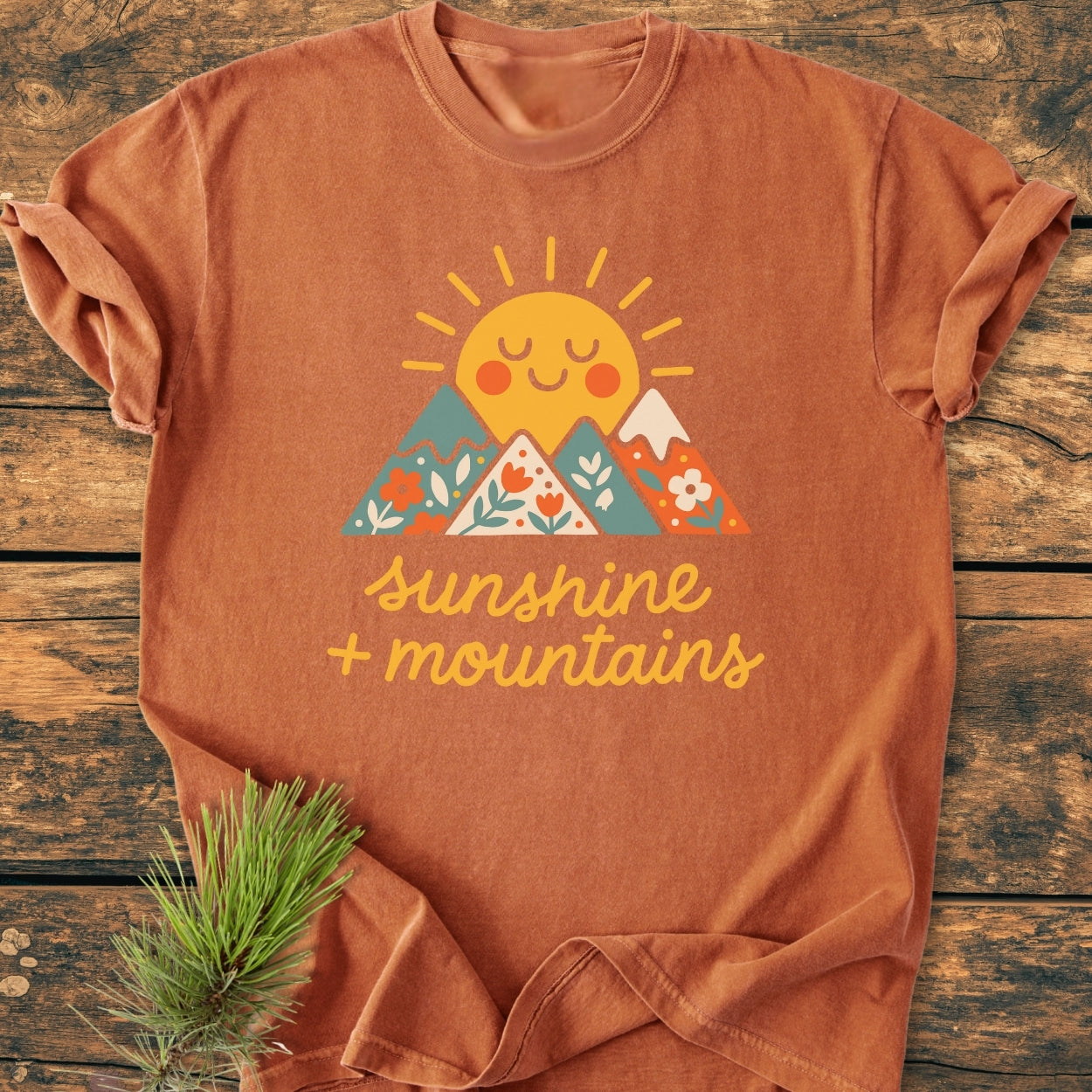 Sunshine And Mountains - Vintage Wash Tee featuring a cartoon sun and flower-patterned mountains design on a rust-orange T-shirt, laid on a wooden surface with a pine sprig beside it.