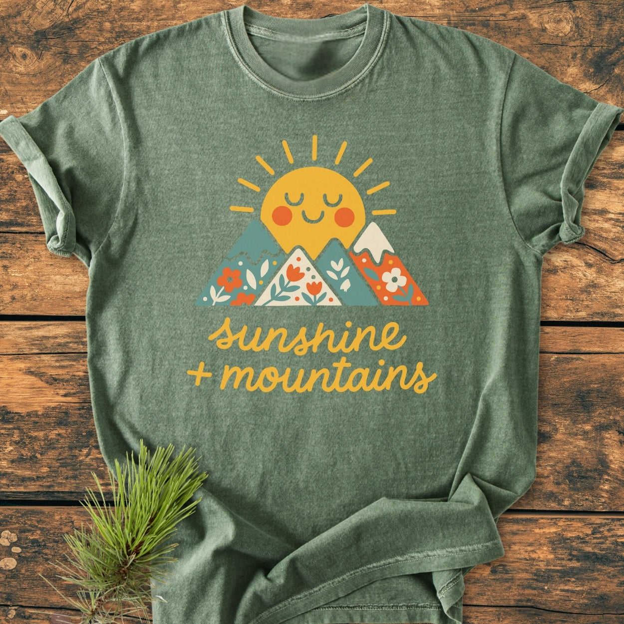 Sunshine And Mountains - Vintage Wash Tee, featuring a smiling sun and floral-patterned mountains graphic, laid on wood. Includes pine sprig accent. Made from pre-shrunk, ring-spun US cotton for a relaxed fit.