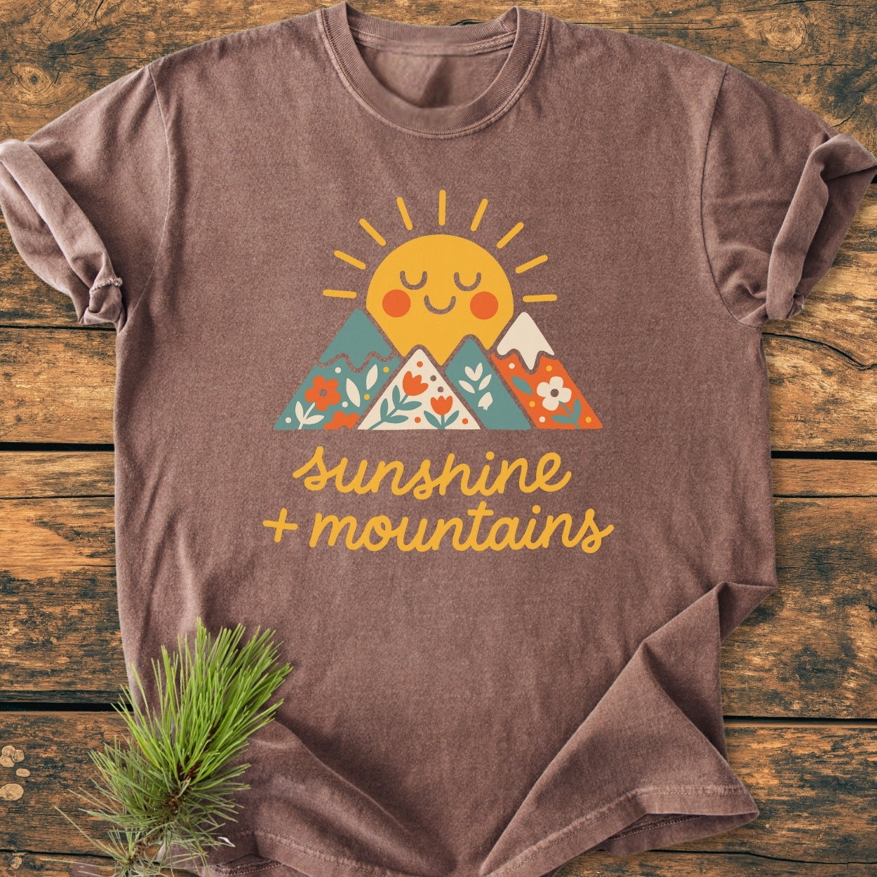 Sunshine And Mountains - Vintage Wash Tee featuring a smiling sun and mountain graphic, laid on wood. Hand-lettered slogan below design. Soft, relaxed fit, perfect for daily wear. Pine sprig accent nearby.