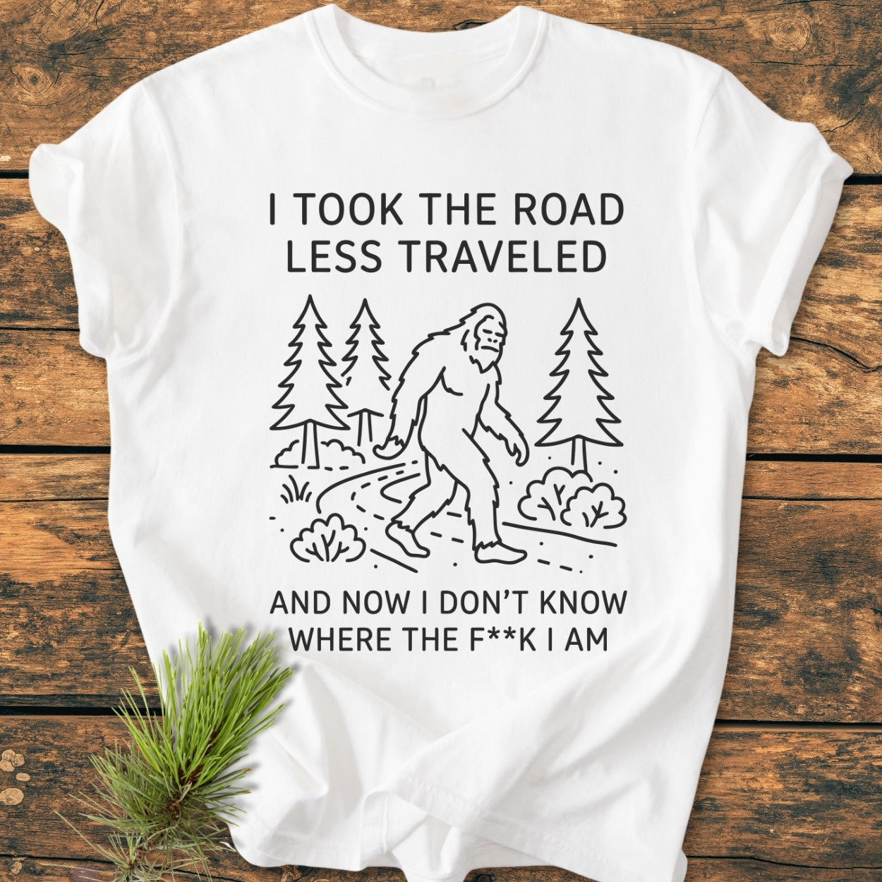 I Took The Road Less Traveled - Vintage Wash Tee: White T-shirt with Bigfoot line drawing and humorous text, laid on wood. Pine sprig accentuates the soft, relaxed garment-dyed cotton design.