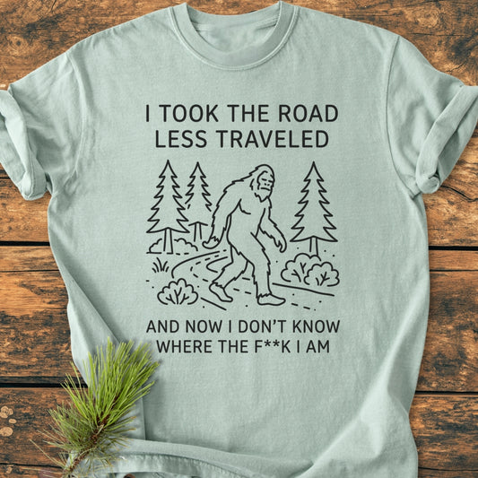 I Took The Road Less Traveled - Vintage Wash Tee features a cartoon Sasquatch on a forest path, with humorous text. Laid on wood with a pine sprig, showcasing its relaxed, garment-dyed design.