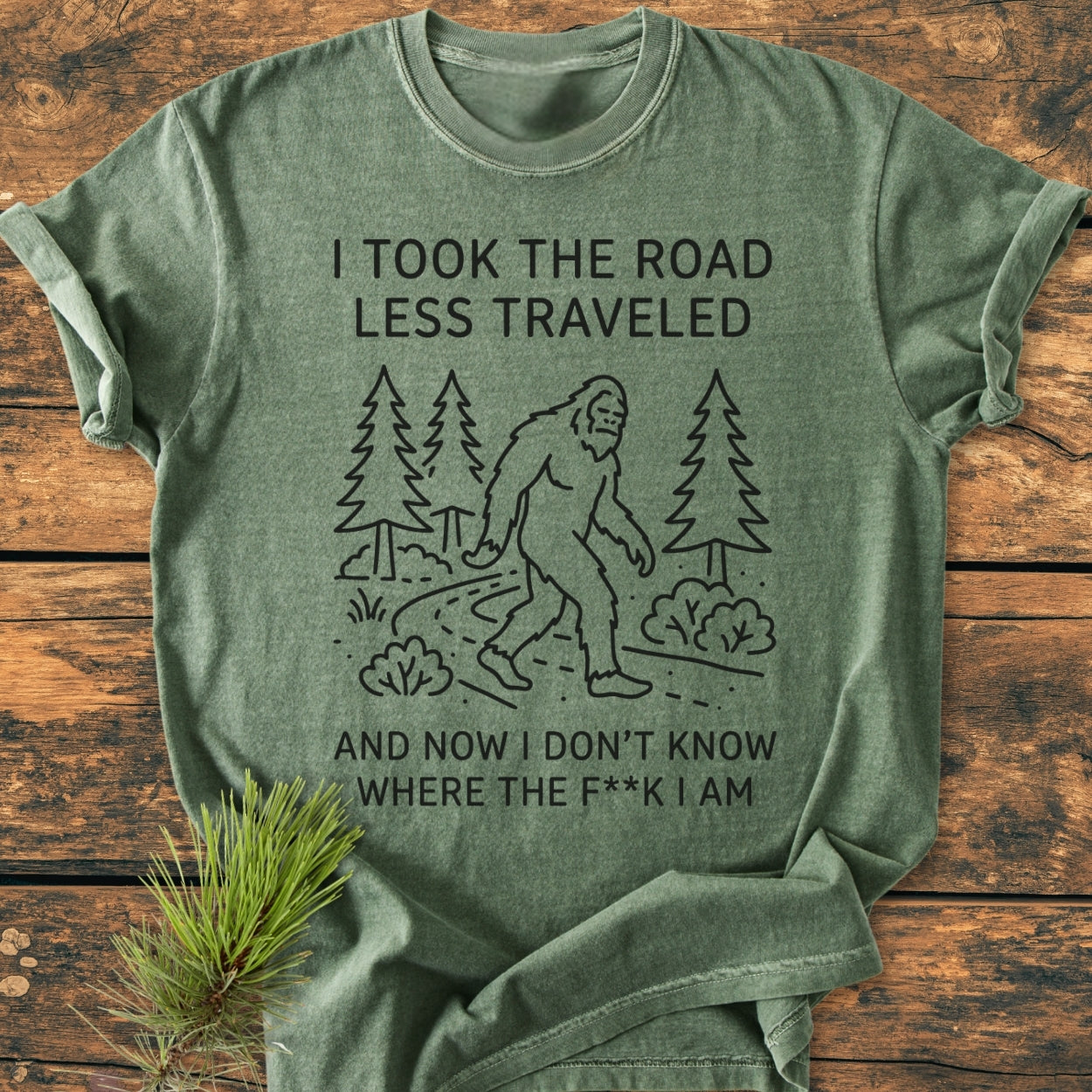 I Took The Road Less Traveled - Vintage Wash Tee features a Bigfoot graphic on a heather-green T-shirt, laid on wood with a pine sprig, enhancing its rustic, adventurous theme.