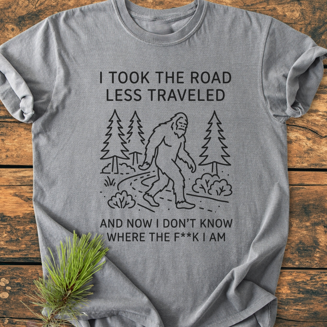 I Took The Road Less Traveled - Vintage Wash Tee features Bigfoot on a forest trail with humorous text. Gray, relaxed fit, 100% cotton, perfect for daily comfort and style.