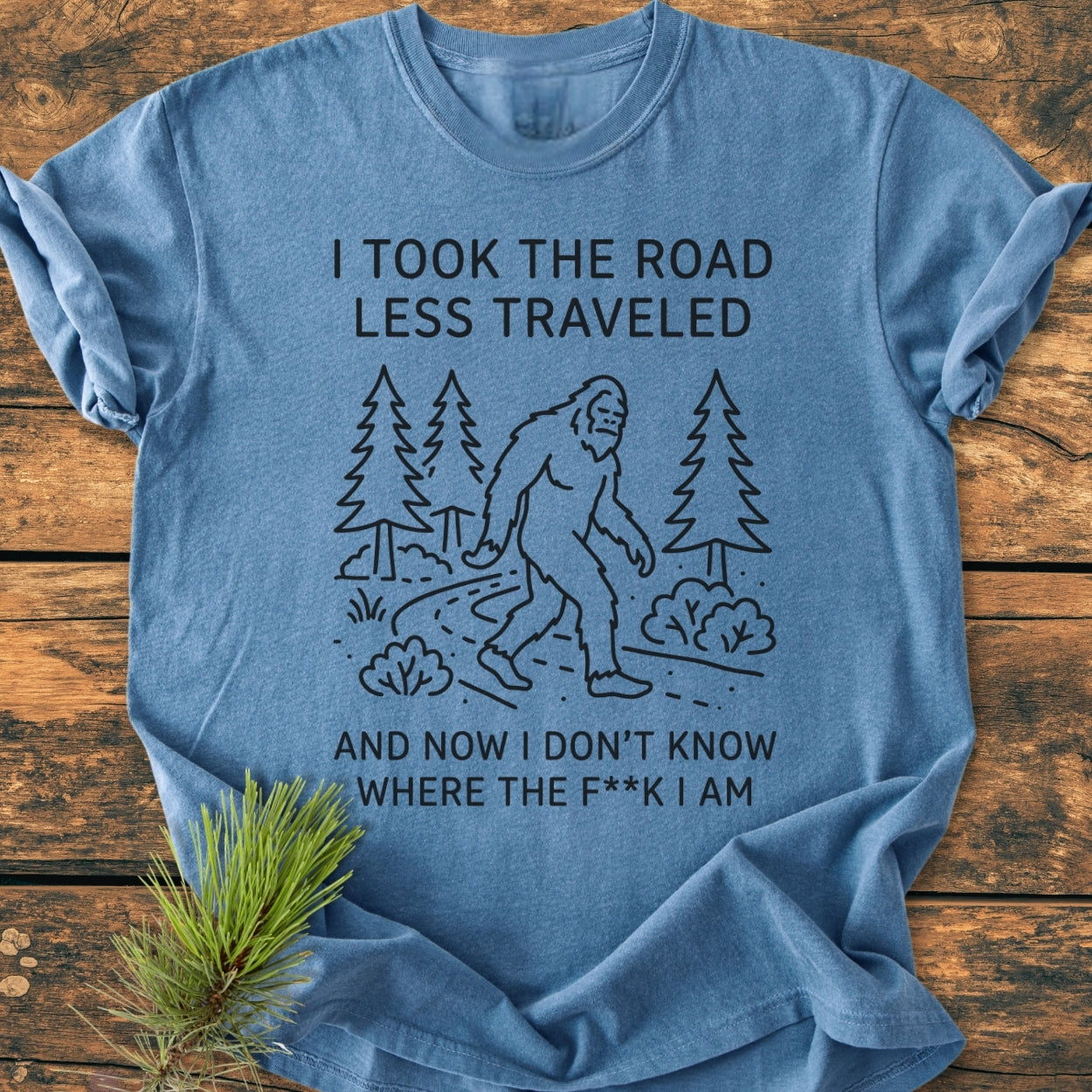 I Took The Road Less Traveled - Vintage Wash Tee featuring Bigfoot on a forest path, printed on a soft, relaxed-fit blue tee laid on a wooden surface with a pine sprig accent.