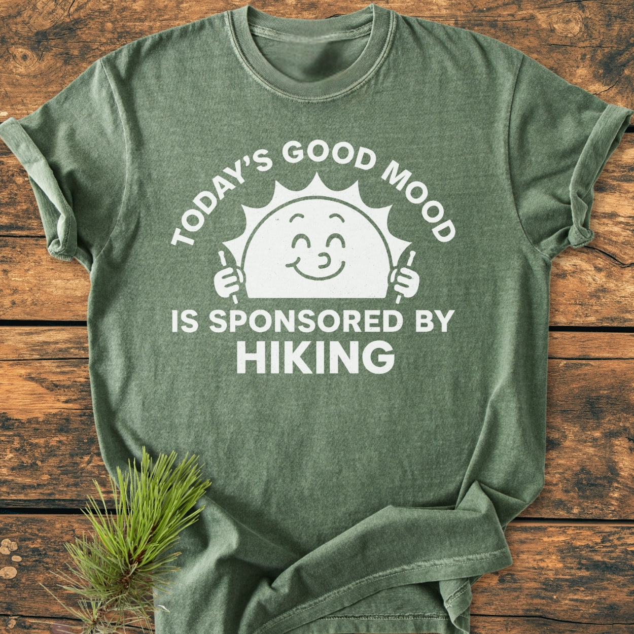 Heather-green Today's Good Mood - Vintage Wash Tee with a smiling sun graphic and hiking message on a wooden surface, accompanied by a sprig of pine needles.