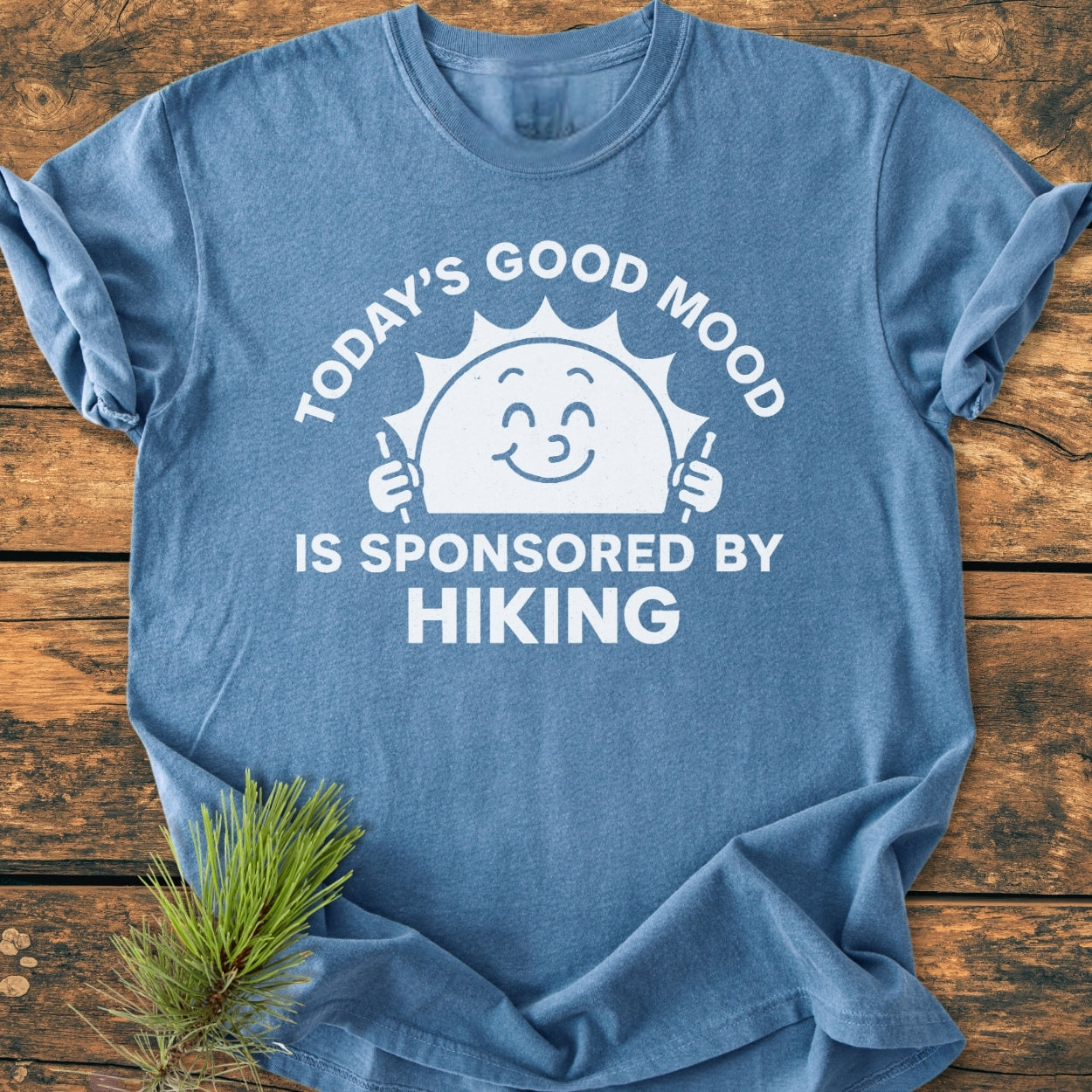 Today's Good Mood - Vintage Wash Tee features a smiling sun with hiking poles and text TODAY’S GOOD MOOD IS SPONSORED BY HIKING on soft, heather-blue fabric, laid on wood.