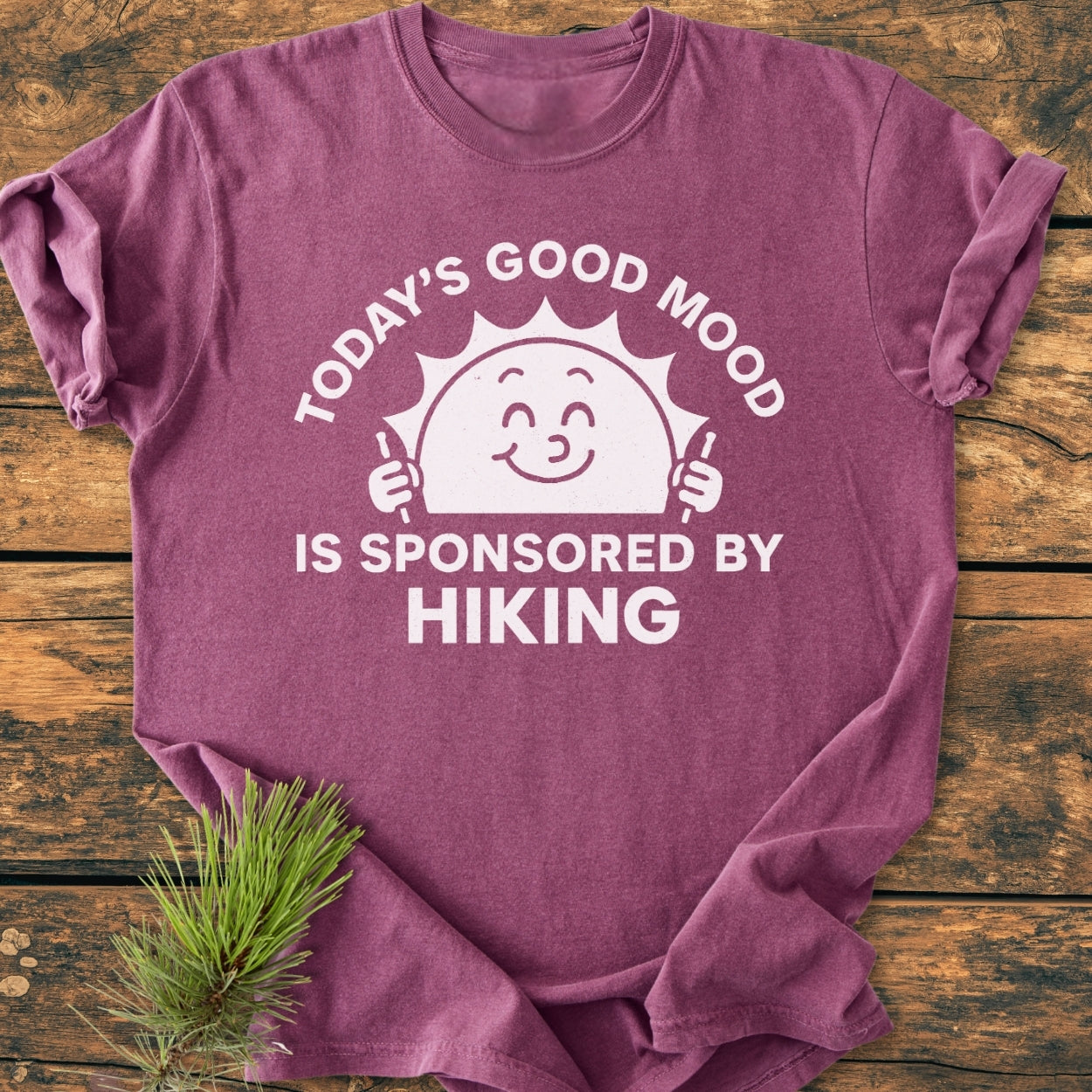 Today's Good Mood - Vintage Wash Tee in heather-purple, features a white graphic of a smiling sun with hiking poles and the slogan about hiking, displayed on a rustic wooden surface with a pine sprig.