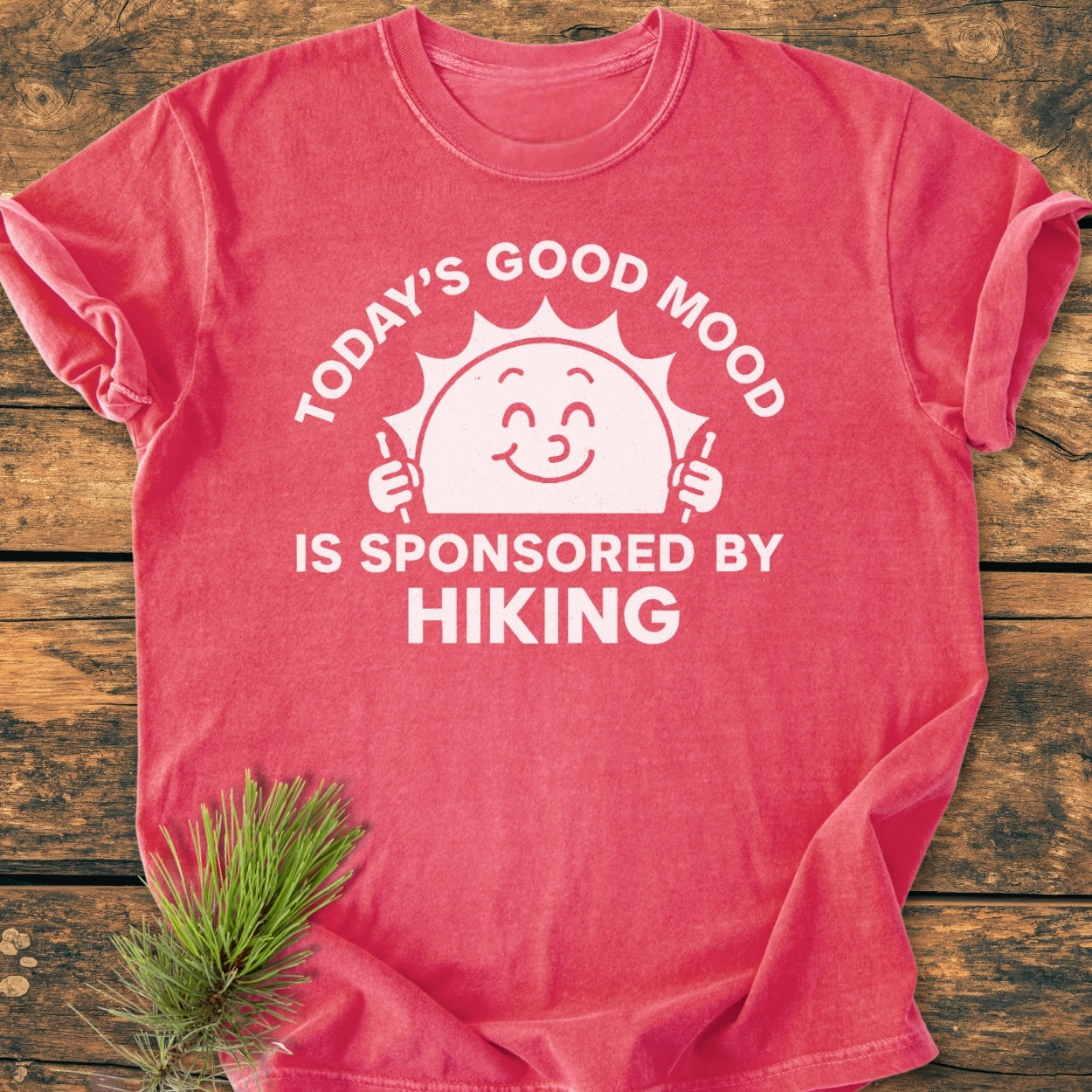 Today's Good Mood - Vintage Wash Tee features a smiling sun with hiking poles and the slogan, “TODAY’S GOOD MOOD IS SPONSORED BY HIKING,” on a flat-laid red-pink shirt.
