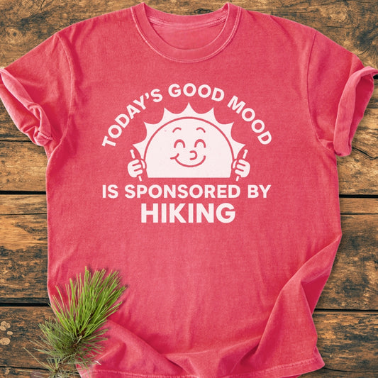 Today's Good Mood - Vintage Wash Tee features a smiling sun with hiking poles and the slogan, “TODAY’S GOOD MOOD IS SPONSORED BY HIKING,” on a flat-laid red-pink shirt.