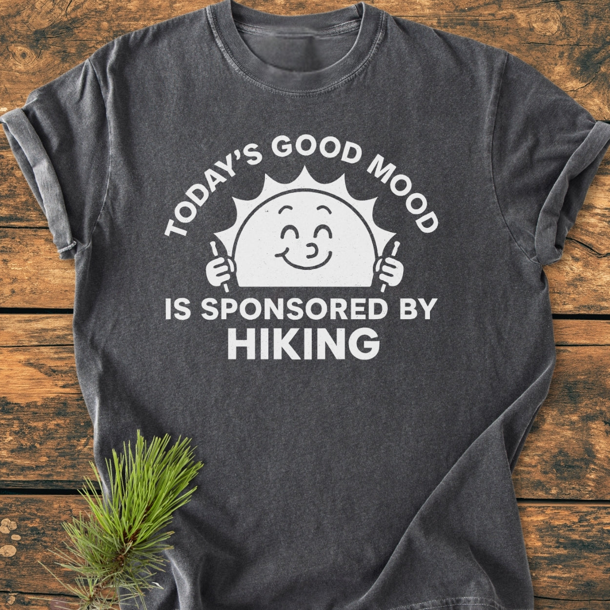 Today's Good Mood - Vintage Wash Tee: Charcoal-gray T-shirt with a smiling sun holding hiking poles and text. Laid flat on wood, styled with a pine sprig for a cozy, outdoor-inspired look.