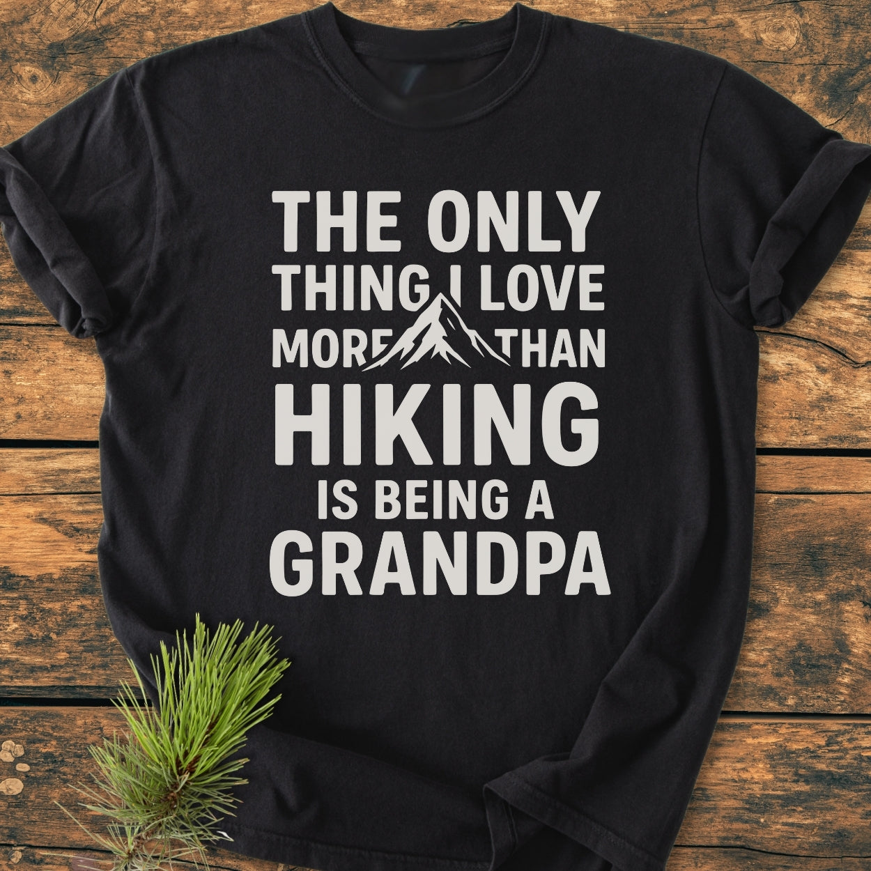 Hiking Grandpa - Vintage Wash Tee: Black T-shirt with bold white text highlighting hiking and grandparenting, featuring a mountain graphic. Laid on wood with a pine branch accentuating the outdoorsy theme.