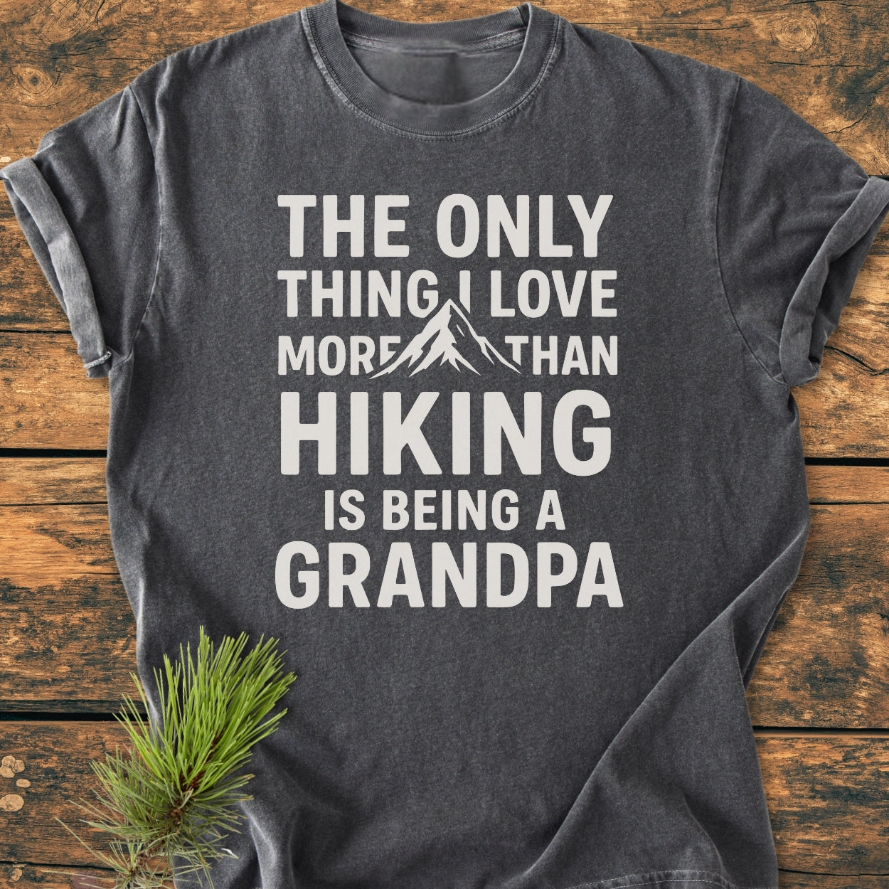 Hiking Grandpa - Vintage Wash Tee, a dark gray T-shirt with a white slogan about hiking and grandparenting, features a mountain graphic. Styled with a pine sprig on a wooden surface.
