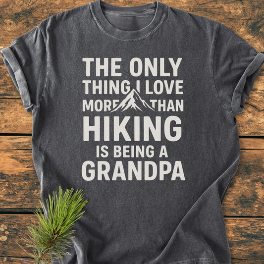 Hiking Grandpa - Vintage Wash Tee, a dark gray T-shirt with a white slogan about hiking and grandparenting, features a mountain graphic. Styled with a pine sprig on a wooden surface.
