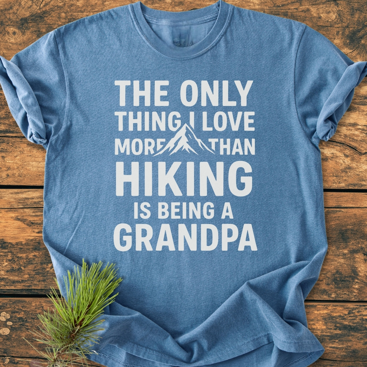 Heather-blue Hiking Grandpa - Vintage Wash Tee with bold white text and mountain graphic, laid on rustic wood with pine branch. Soft, garment-dyed cotton, relaxed fit, and durable stitching for comfort.
