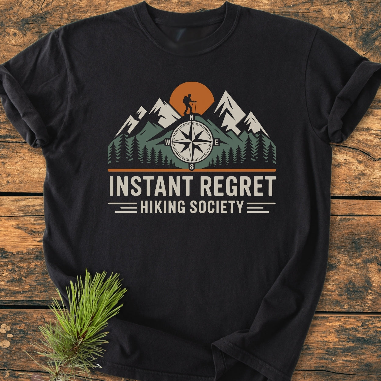 Instant Regret Hiking Society - Vintage Wash Tee featuring a stylized mountain scene with a hiker and compass rose, laid on wood, highlighting its soft-washed, durable 100% cotton fabric.