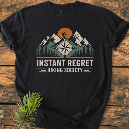 Instant Regret Hiking Society - Vintage Wash Tee featuring a stylized mountain scene with a hiker and compass rose, laid on wood, highlighting its soft-washed, durable 100% cotton fabric.