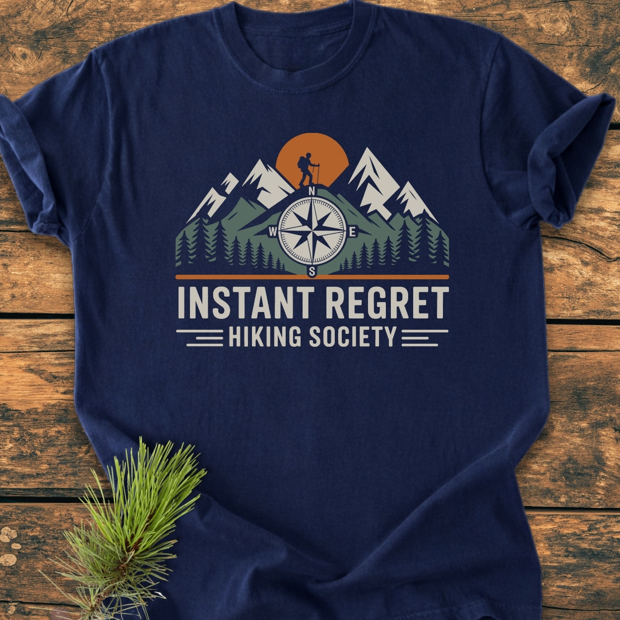 Instant Regret Hiking Society - Vintage Wash Tee features mountains, pine trees, a compass rose, and a hiker silhouette against an orange sun on navy-blue fabric, laid on a wooden surface.
