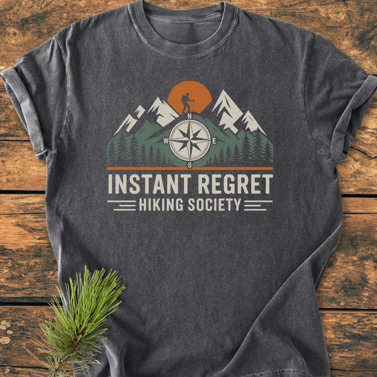 Instant Regret Hiking Society - Vintage Wash Tee features a mountain and hiker graphic with a compass and sun, showcased on a charcoal-gray T-shirt, laid flat with a pine sprig accent.