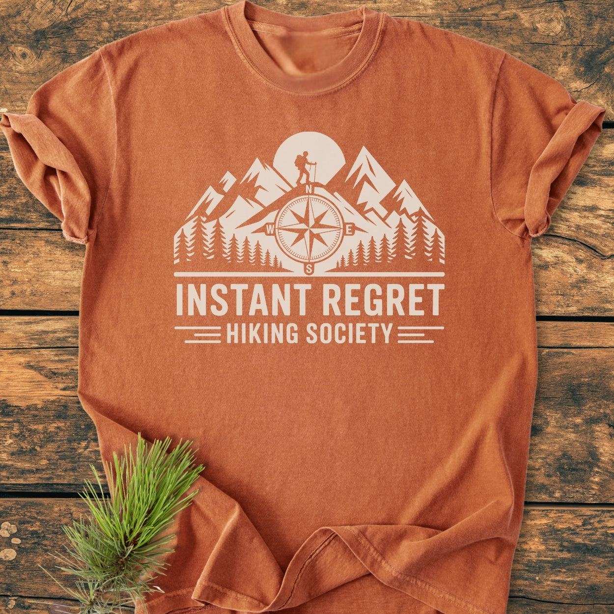 Instant Regret Hiking Society - Vintage Wash Tee features a stylized mountain and hiker design on a rust-orange T-shirt with a cozy, relaxed fit, lying on a wooden surface.