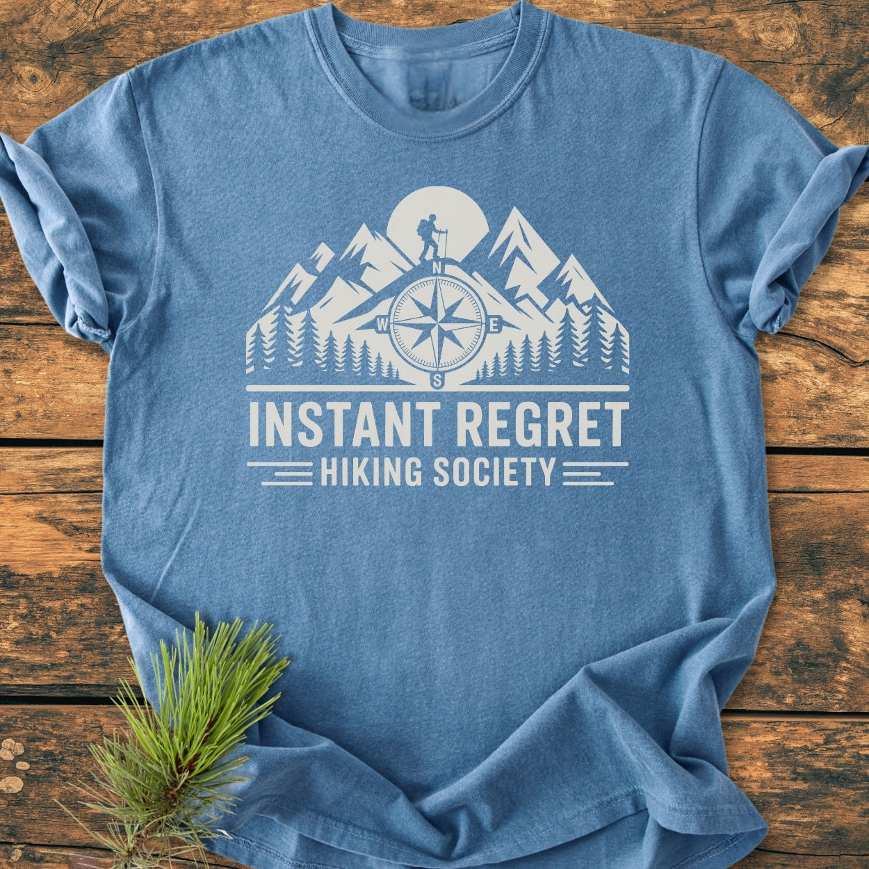 Instant Regret Hiking Society - Vintage Wash Tee features a stylized outdoor scene with mountains and a hiker, printed on a heather-blue Comfort Colors T-shirt, laid flat on a wooden surface.