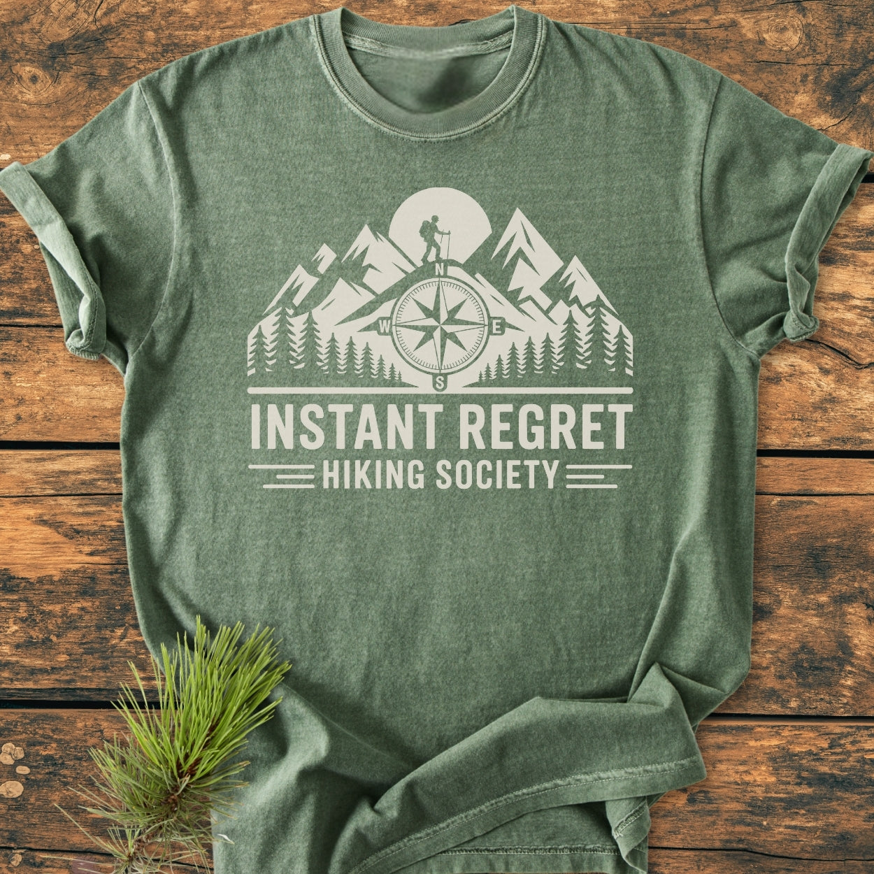 Instant Regret Hiking Society - Vintage Wash Tee in heather-green with off-white outdoor scene graphic and slogan, laid flat on a wooden surface with pine sprig; shows relaxed fit and durable stitching.