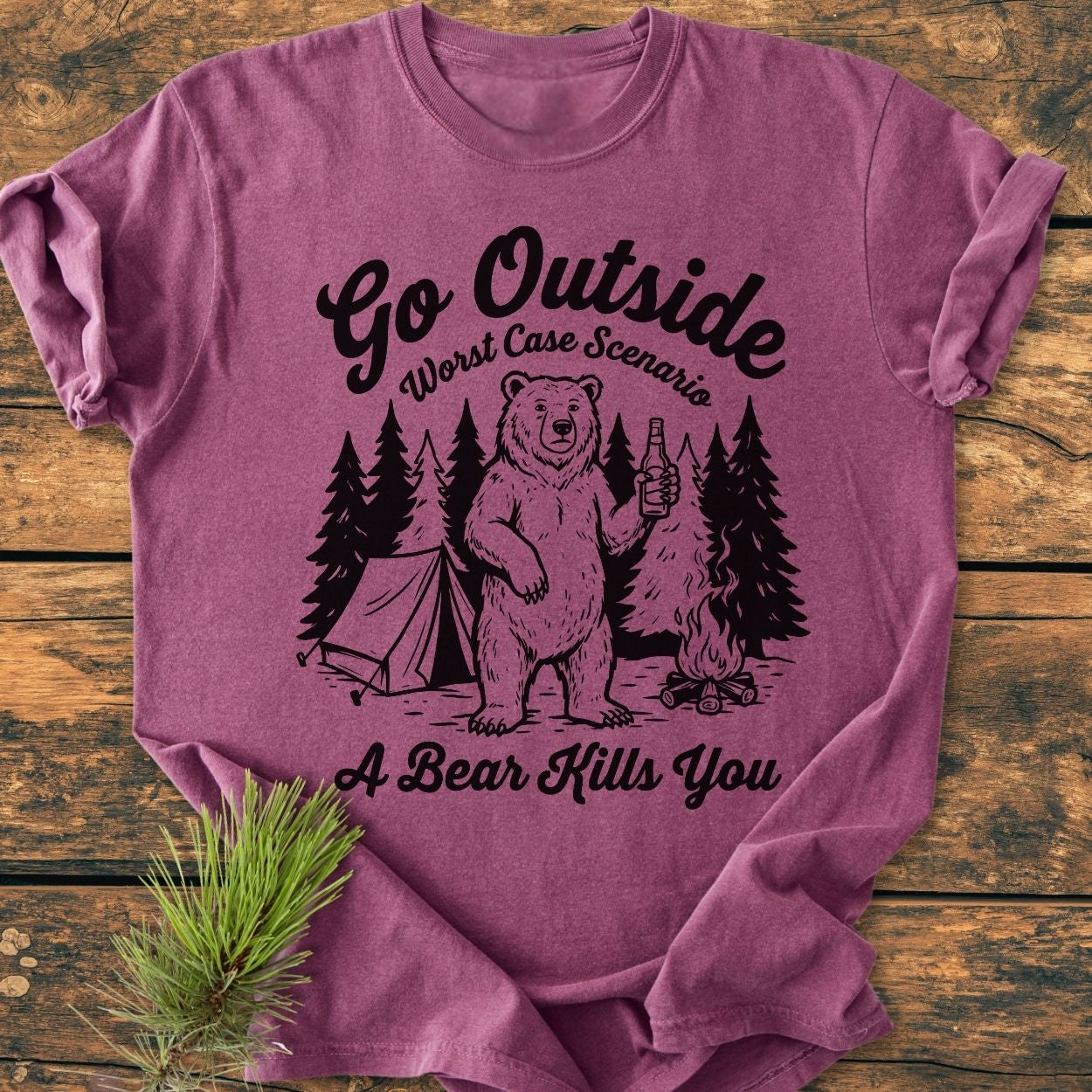 Go Outside - Vintage Wash Tee featuring a bear camping graphic with a tent, trees, and campfire, laid out flat. Nearby, a pine sprig emphasizes the outdoor theme. Made of soft, durable ring-spun cotton.