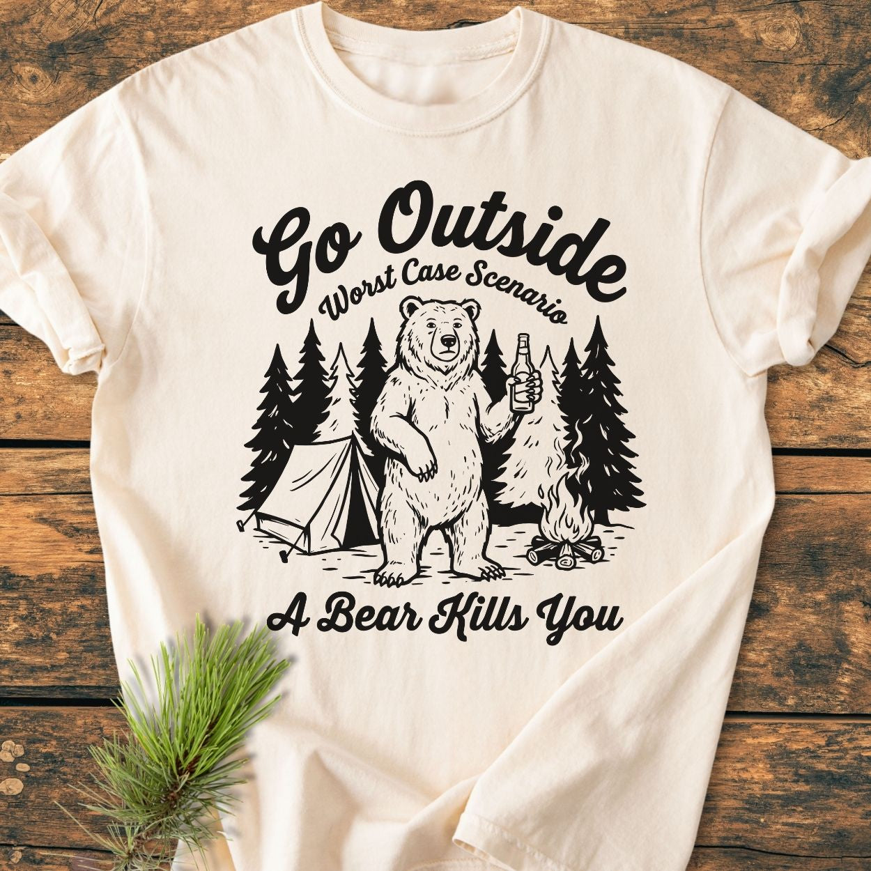 Go Outside - Vintage Wash Tee features a black line-drawing of a whimsical forest camp scene with a bear, tent, campfire, and slogan, printed on a cream-colored T-shirt laid on a wooden surface.
