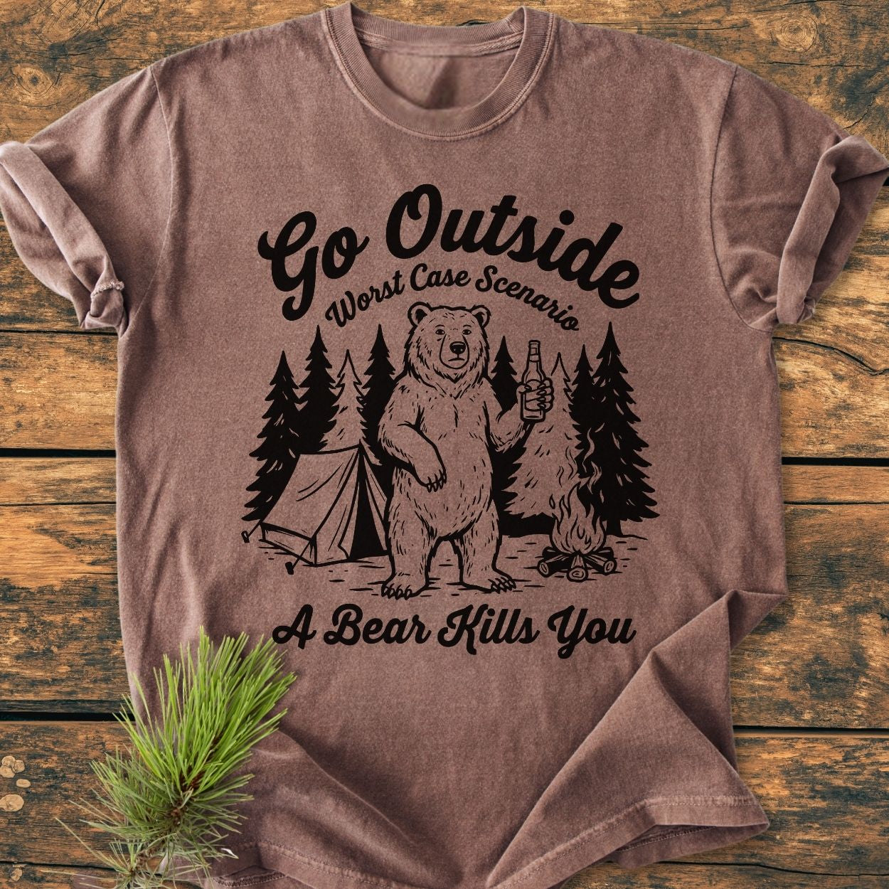 Go Outside - Vintage Wash Tee features a black line-art camping scene with a cartoon bear on a heather-brown T-shirt, laid on a rustic wooden surface with a pine sprig for styling.