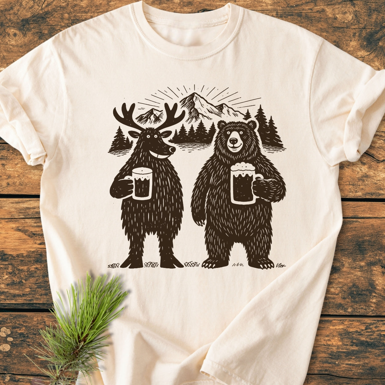 Bear, Deer And Beer - Vintage Wash Tee features playful moose and bear with beer mugs, set against pine trees and mountain backdrop, enhancing its rustic charm on soft, relaxed-fit, cream-colored fabric.