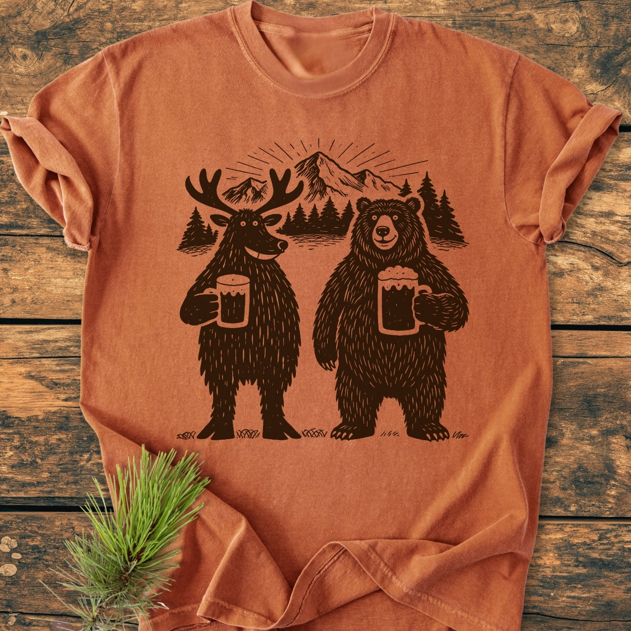Bear, Deer And Beer - Vintage Wash Tee features a moose and bear with beer mugs, mountain backdrop, soft-washed, garment-dyed cotton for comfort, laid flat with a pine sprig accent.
