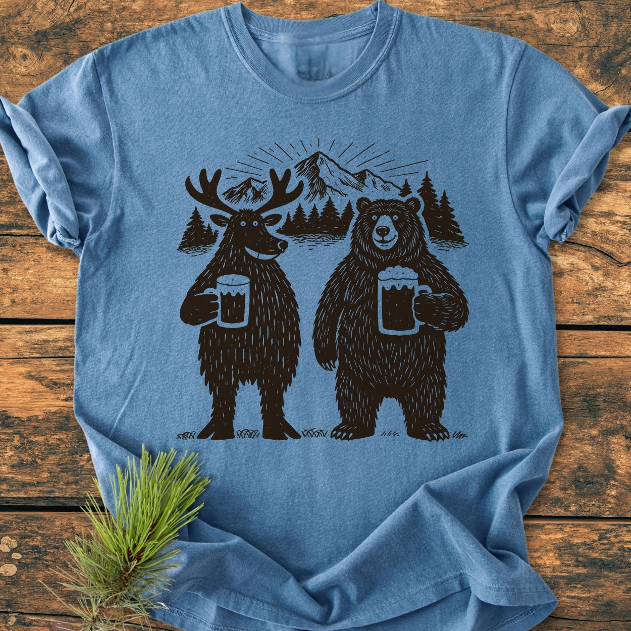 Bear, Deer And Beer - Vintage Wash Tee: Heather-blue T-shirt featuring a whimsical moose and bear with beer mugs, set against pine trees and mountain silhouette. Laid on wooden surface with a pine sprig.