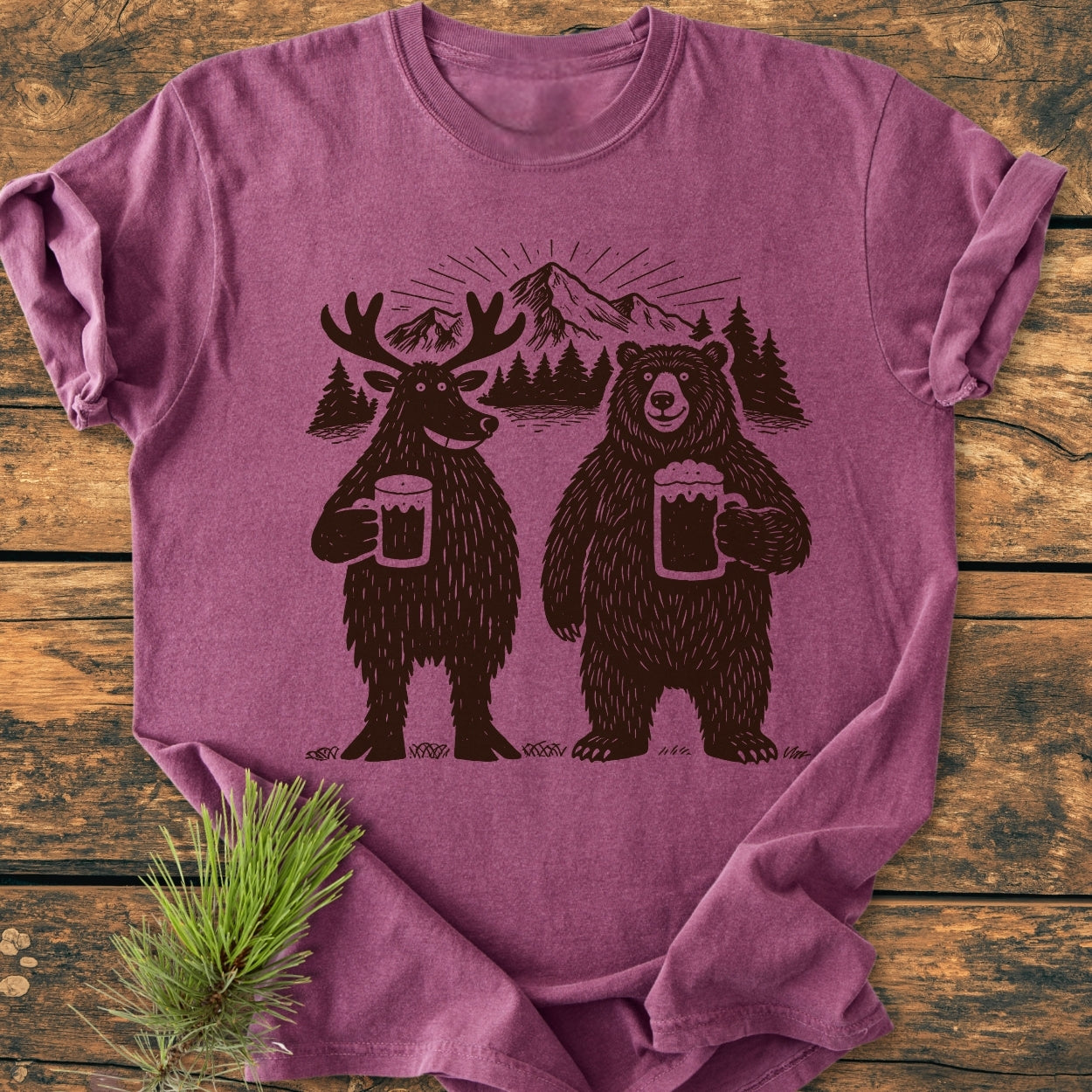 Bear, Deer And Beer - Vintage Wash Tee features a whimsical forest scene with a cartoon moose and bear toasting beer, set against mountains and trees, displayed on a rustic wooden surface.