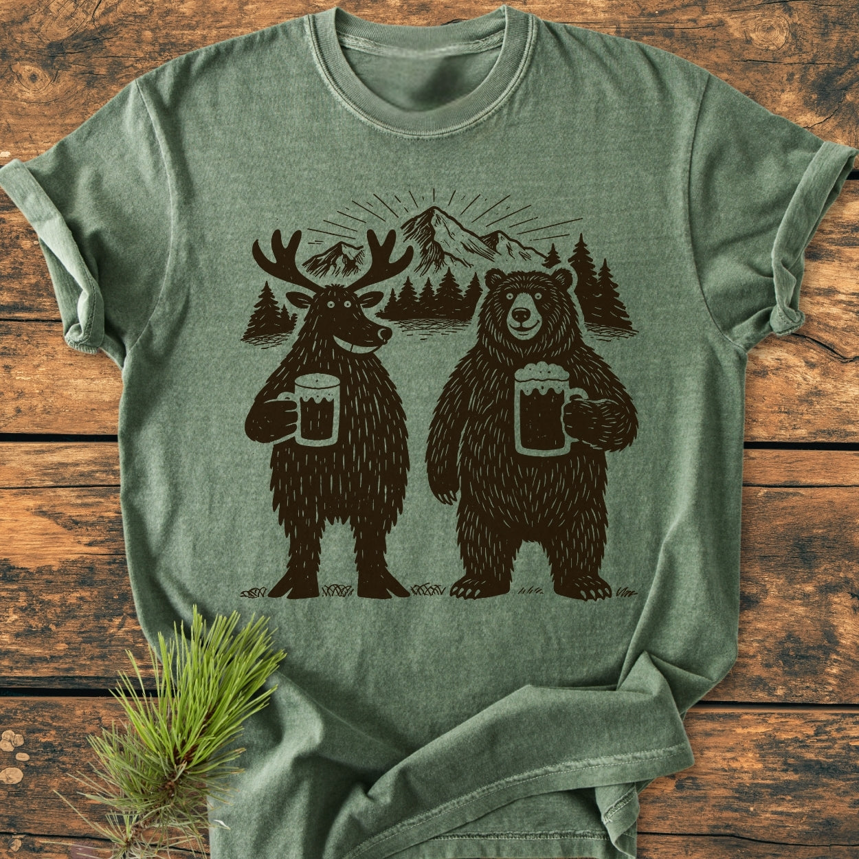 Bear, Deer And Beer - Vintage Wash Tee featuring a playful woodland scene with a moose and bear clinking beer mugs, surrounded by pine trees and mountains, styled on a wooden surface with an evergreen sprig.