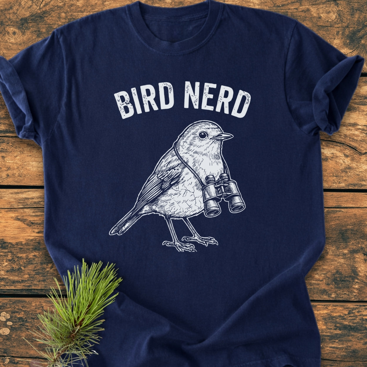 Bird Nerd - Vintage Wash Tee features a songbird with binoculars on a navy-blue shirt, laid on wood. Its relaxed fit and soft-washed fabric offer comfort, accentuated by durable double-needle stitching.