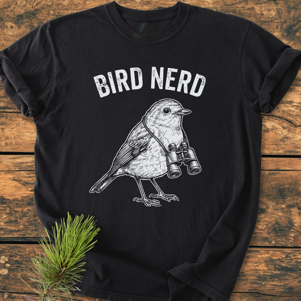 Bird Nerd - Vintage Wash Tee features BIRD NERD text and a songbird with binoculars on soft black fabric, styled with a pine sprig on a wooden surface.