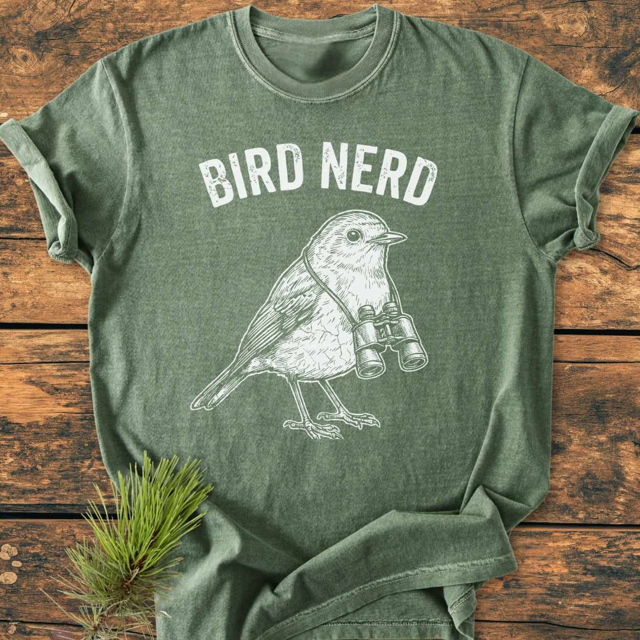 Heather-green Bird Nerd - Vintage Wash Tee featuring a white screen-printed phrase and a line-drawing of a songbird with binoculars, laid on wood with a pine sprig beside it.