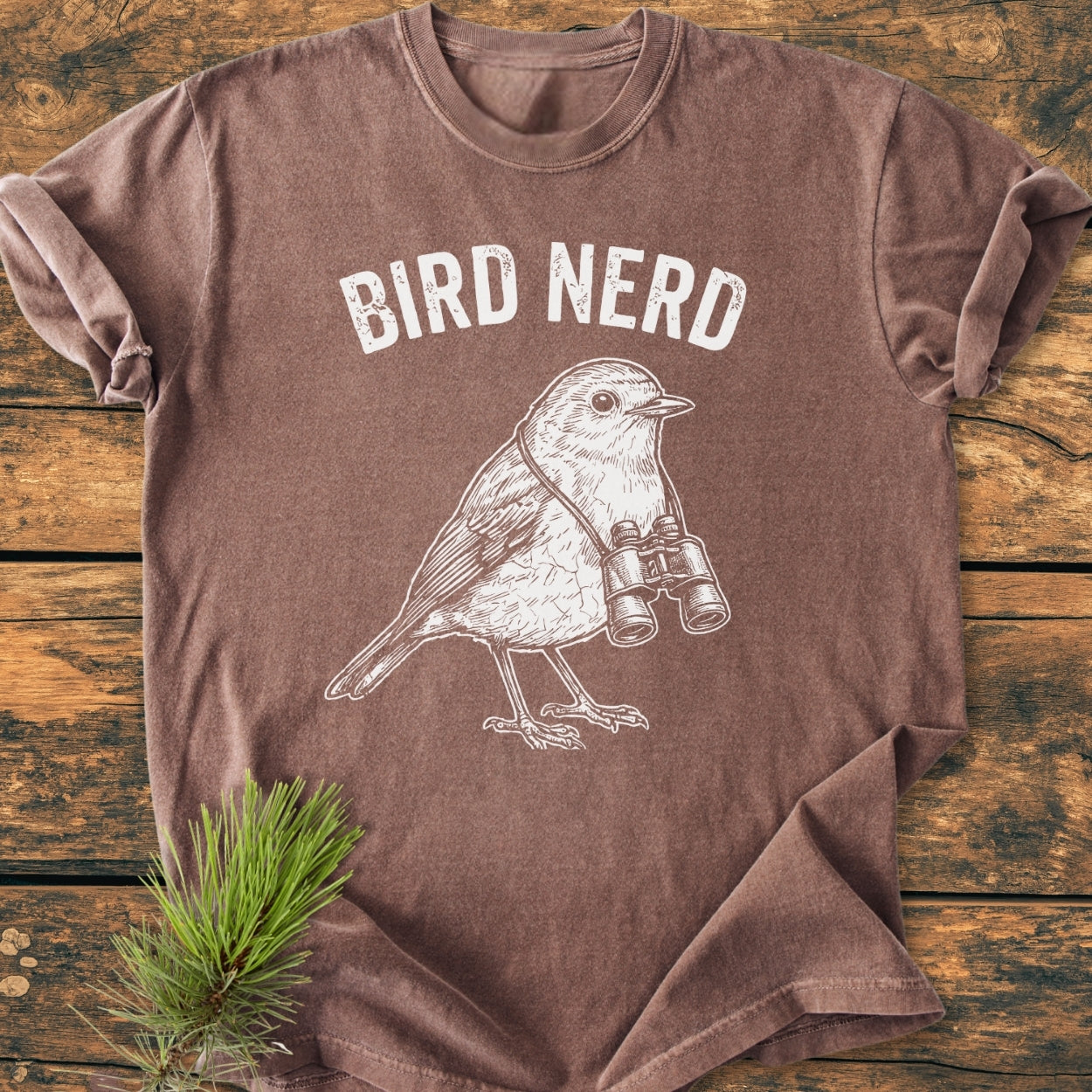 Bird Nerd - Vintage Wash Tee featuring a white line-drawing of a songbird with binoculars and BIRD NERD slogan, laid flat on wood with a pine sprig accent.